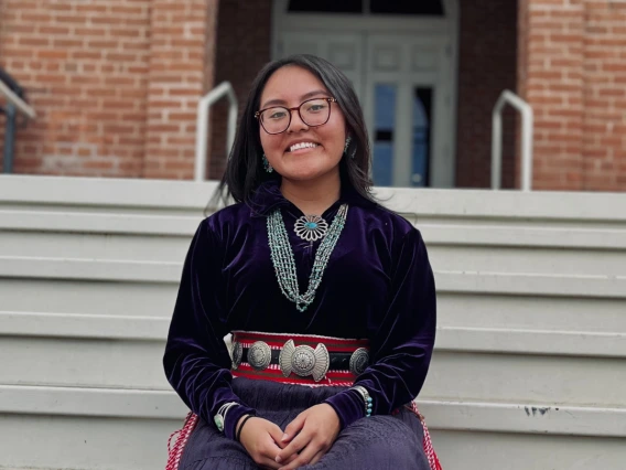 Photo of Kyra Long wearing her traditional regalia, sitting on a staircase.