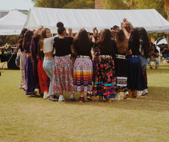 Photo of Indigenous Women holding hands in a circle wearing traditional wear.