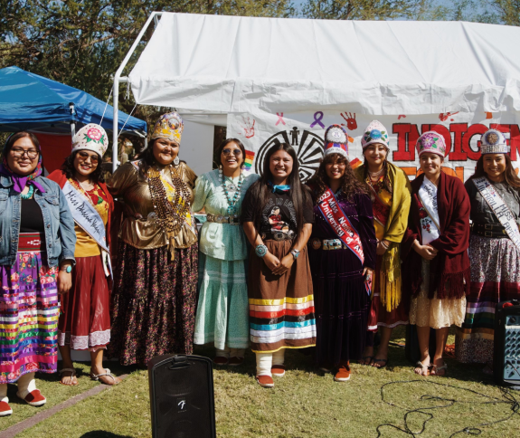 Group photo of Native women in their traditional wear in front of a large banner that reads "Indigenous Peoples Day"
