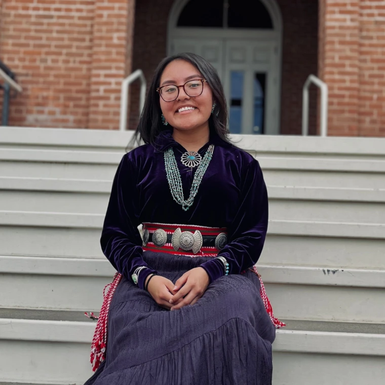 Photo of Kyra Long wearing her traditional regalia, sitting on a staircase.